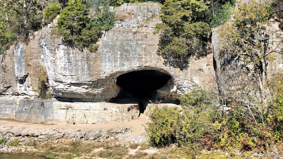 A natural rock formation with a cave entrance, surrounded by trees and a river.