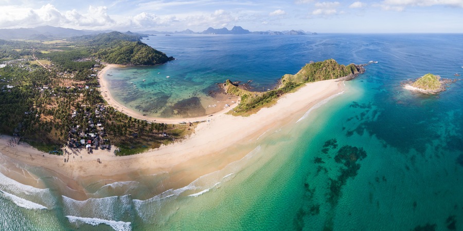 A coastal landscape with a sandy beach, clear turquoise waters, and lush green hills.
