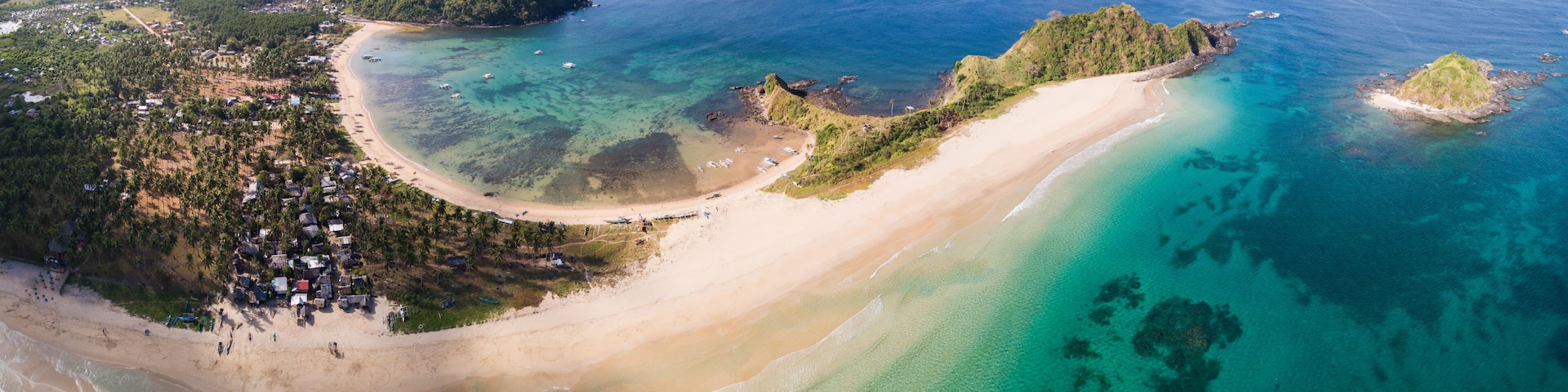 A coastal landscape with a sandy beach, clear turquoise waters, and lush green hills.