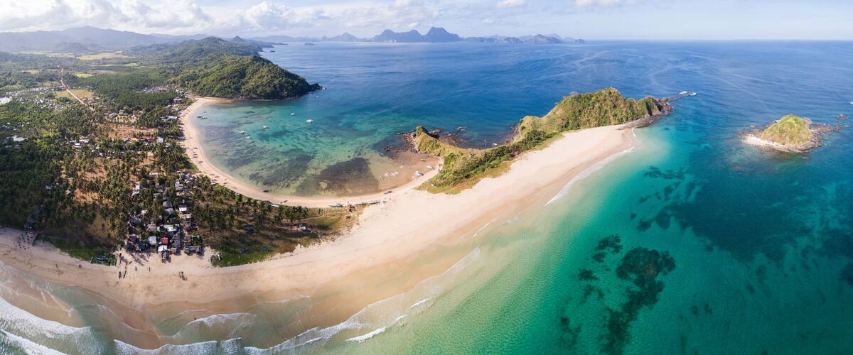A coastal landscape with a sandy beach, clear turquoise waters, and lush green hills.