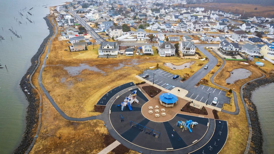 An aerial view of a coastal residential area with houses, a playground, and a parking lot.