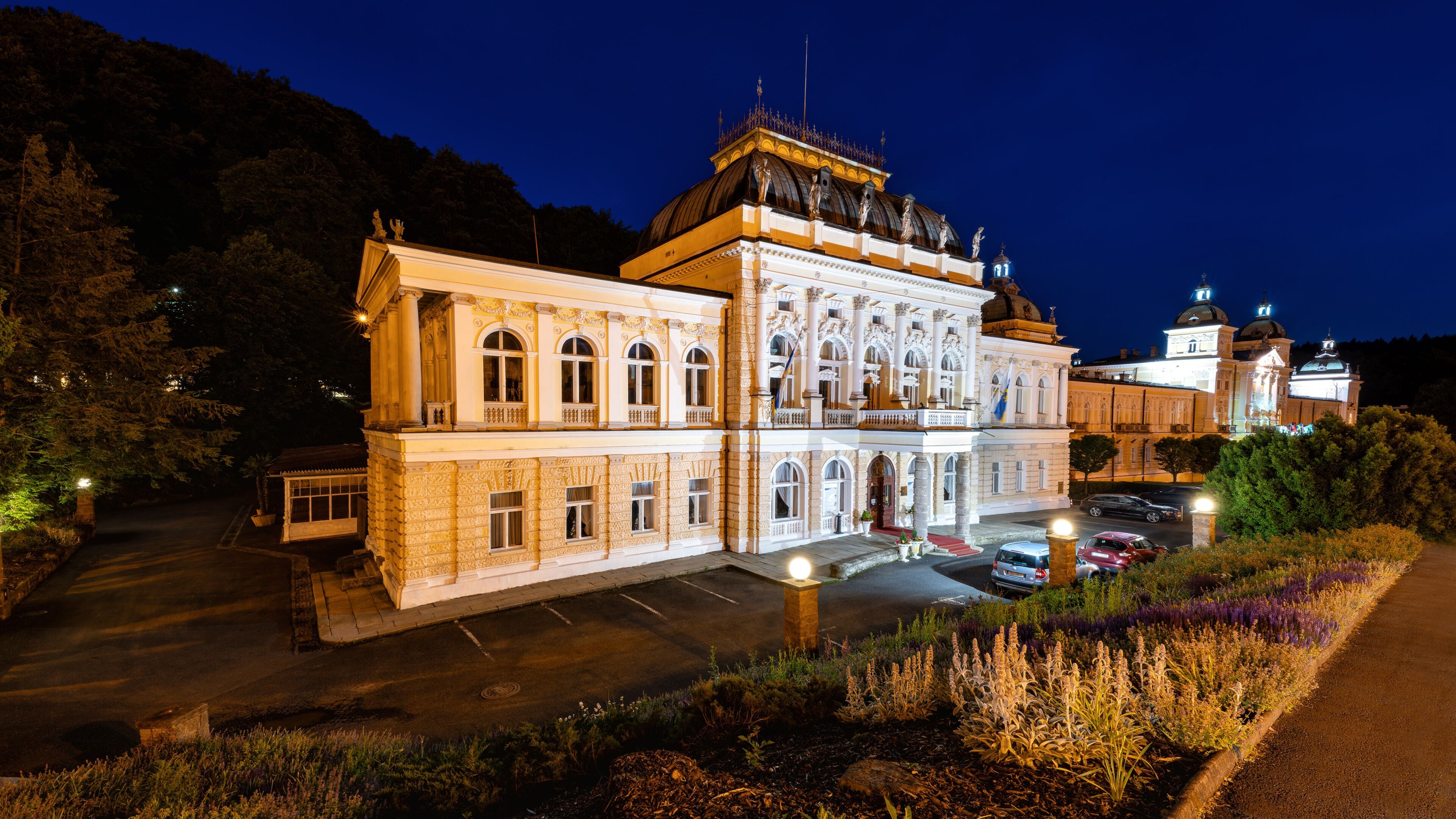 Un edificio imponente e iluminado de arquitectura clásica, rodeado de un jardín ajardinado y coches aparcados.
