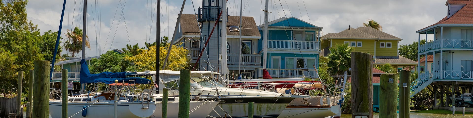 A marina with several boats docked, wooden posts, and colorful houses in the background.