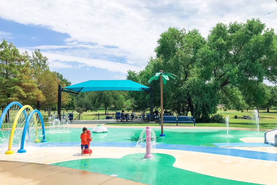 Un parc aquàtic amb una zona de jocs d'aigua, bancs i arbres.