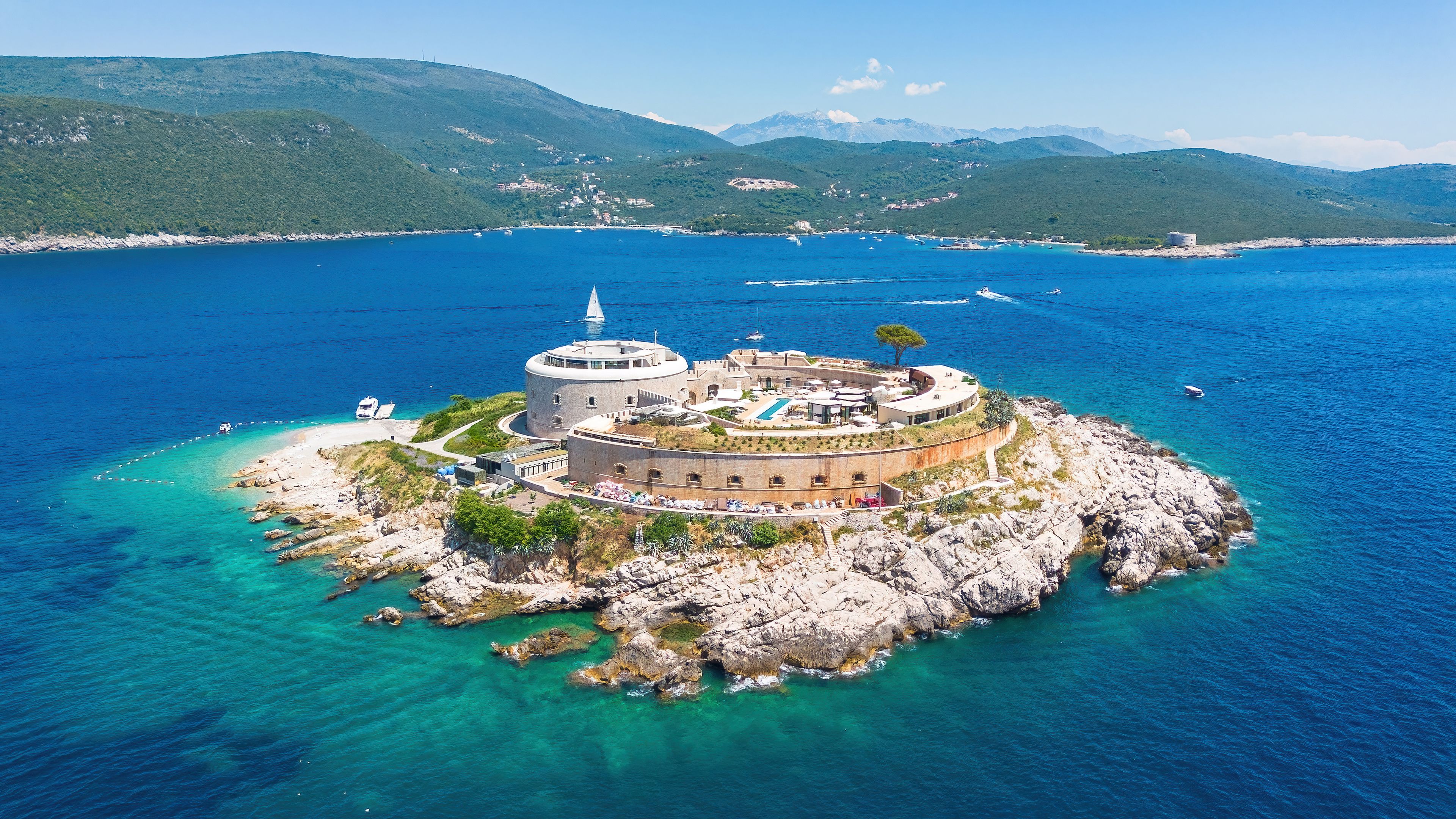 Eine Küsteninsel mit einer historischen Festung, klarem türkisblauem Wasser und einem gebirgigen Hintergrund.