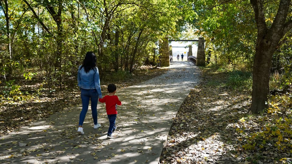 A paved pathway in a park with trees and a stone bridge in the distance.