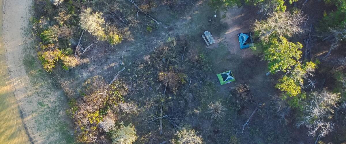 An aerial view of a coastal area with a sandy beach, dense vegetation, and three tents.