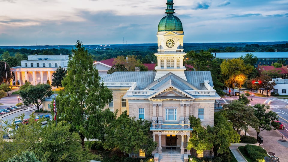 A historic building with a clock tower, surrounded by trees and a cityscape in the background.