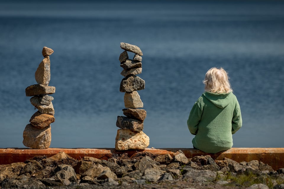 A person sitting on a wooden platform by the sea, overlooking two stone cairns.