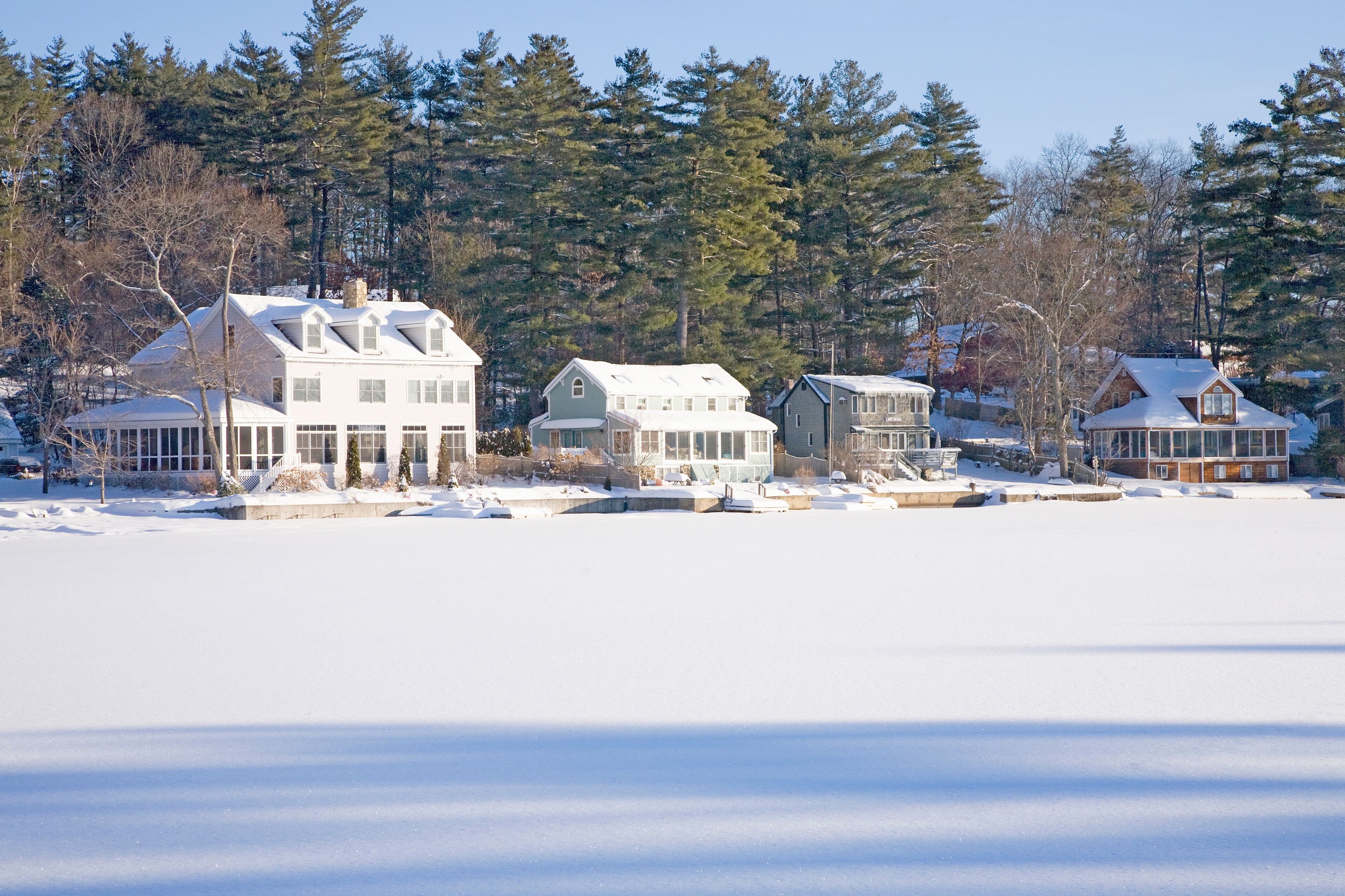 A winter scene with a row of houses by a frozen lake, surrounded by trees.