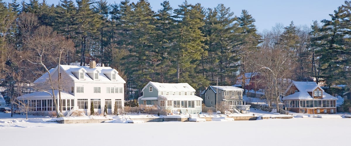 A winter scene with a row of houses by a frozen lake, surrounded by trees.
