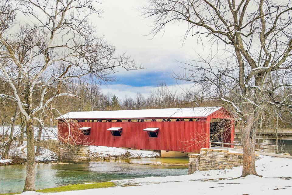 Un puente cubierto de color rojo cruzando un río, con nieve en el suelo y árboles.