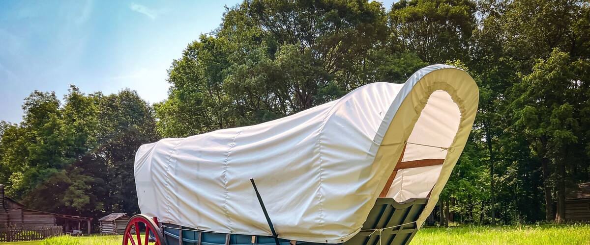 Rear view of a 19th century style horse-drawn carriage or covered wagon set against a green rural nature background.