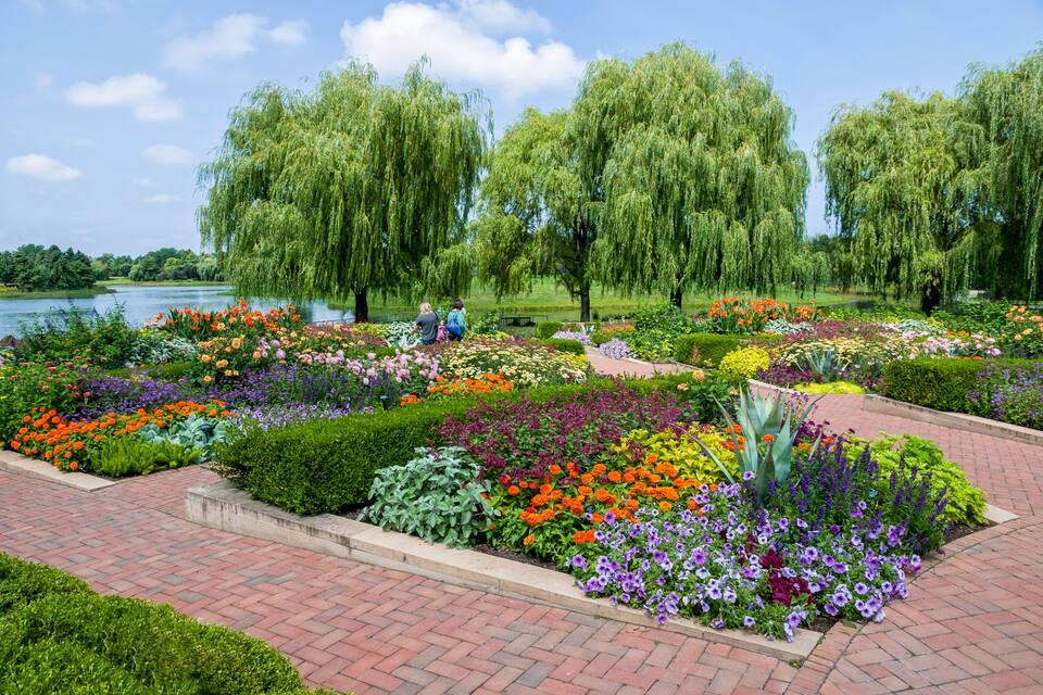 A park with a lake, colorful flower beds, and weeping willow trees.