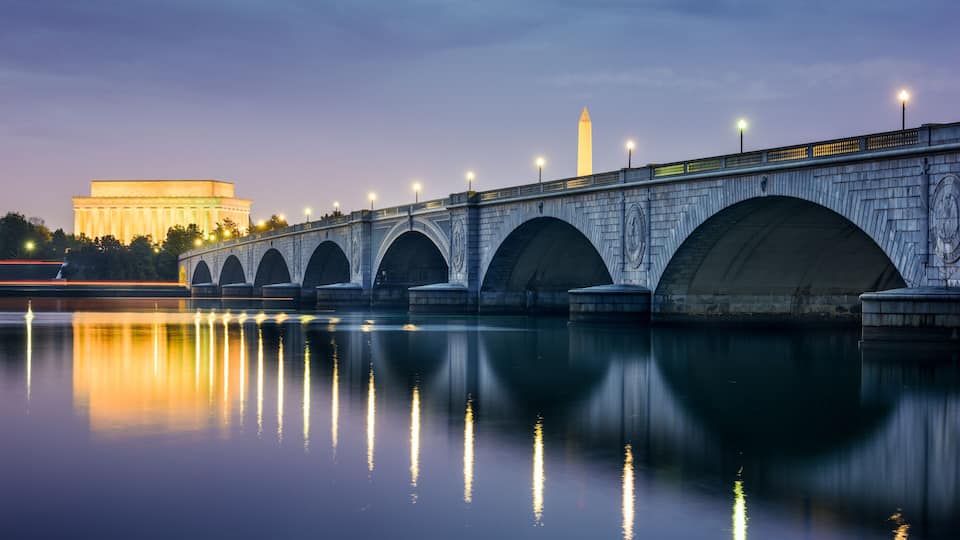 A stone bridge with arches over a calm body of water, reflecting lights and a distant illuminated monument.