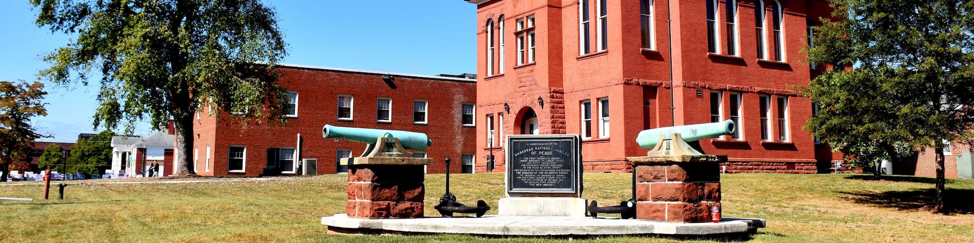 A red brick building with a clock tower, two cannons, and a monument in front.