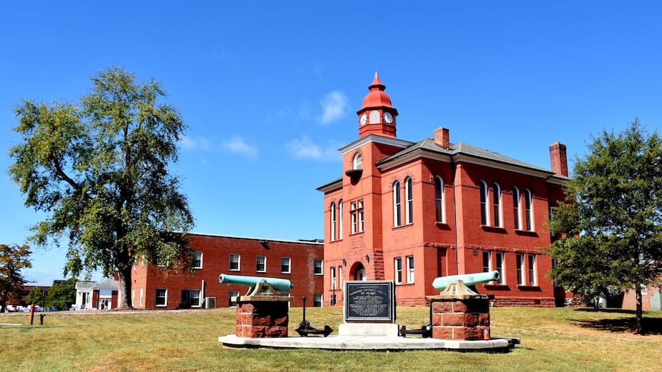 A red brick building with a clock tower, two cannons, and a monument in front.