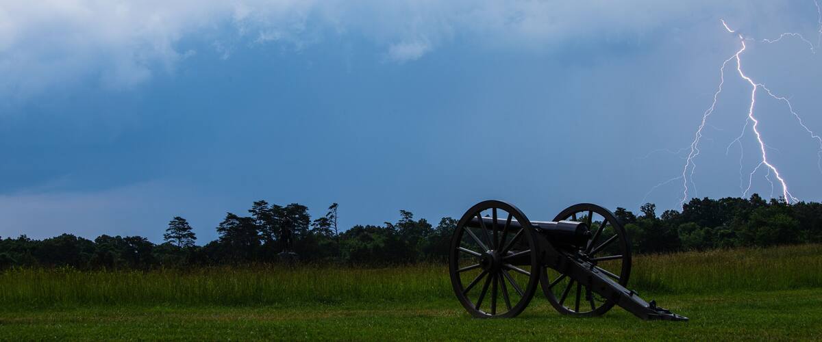 A cannon in a field with a stormy sky and lightning.