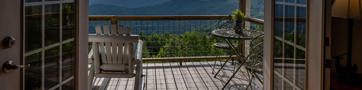 Une terrasse en bois avec une chaise blanche et une petite table, d’où l’on peut admirer une chaîne de montagnes.
