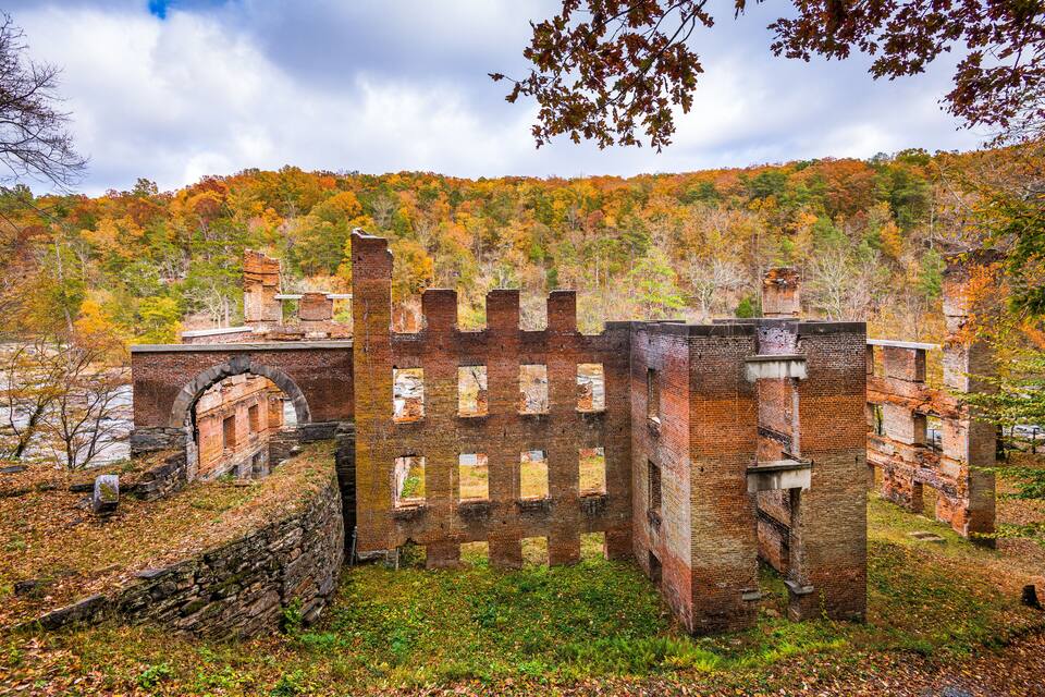 An abandoned brick building with arched windows and a stone wall in a forested area with autumn foliage.