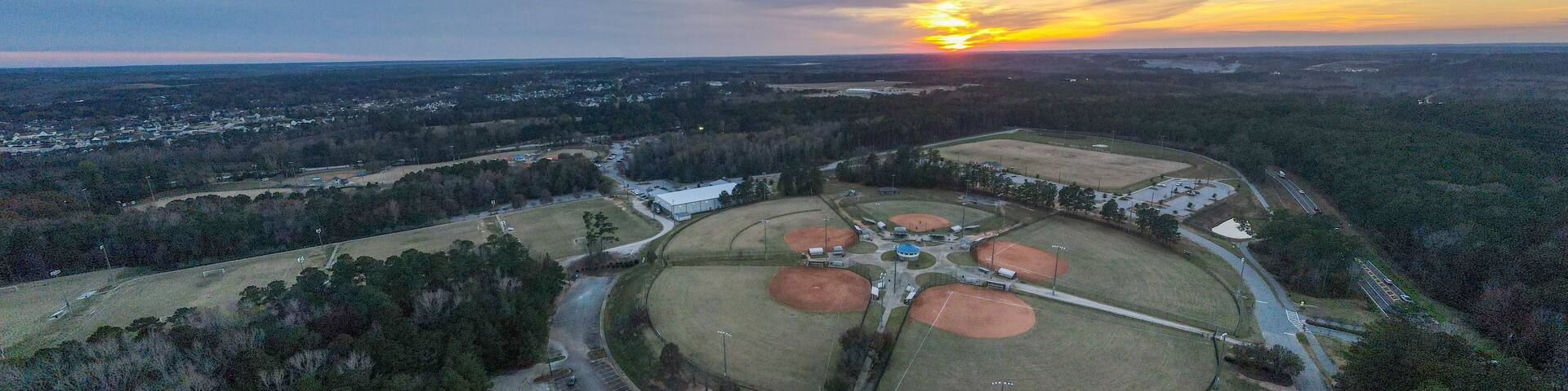An aerial view of a baseball field surrounded by dense forest, with a sunset in the background.