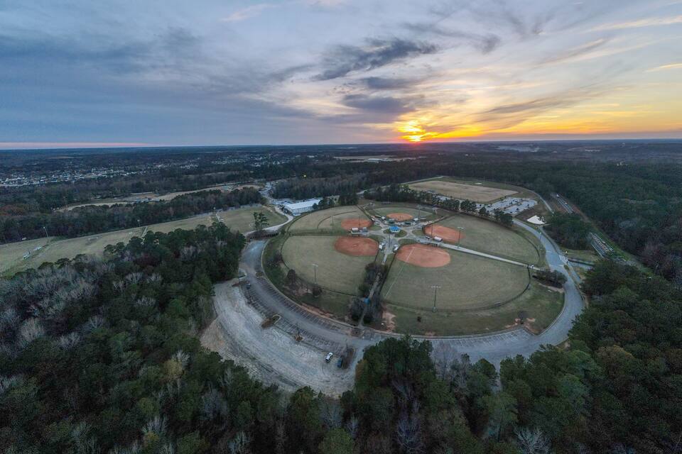 Eine Vogelperspektive auf ein Baseballfeld, umgeben von dichtem Wald, mit einem Sonnenuntergang im Hintergrund.