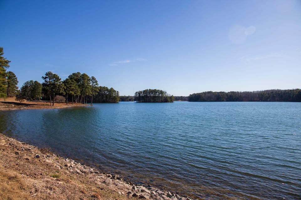 Um lago tranquilo com uma pequena ilha ao centro, rodeado por árvores e um céu limpo.