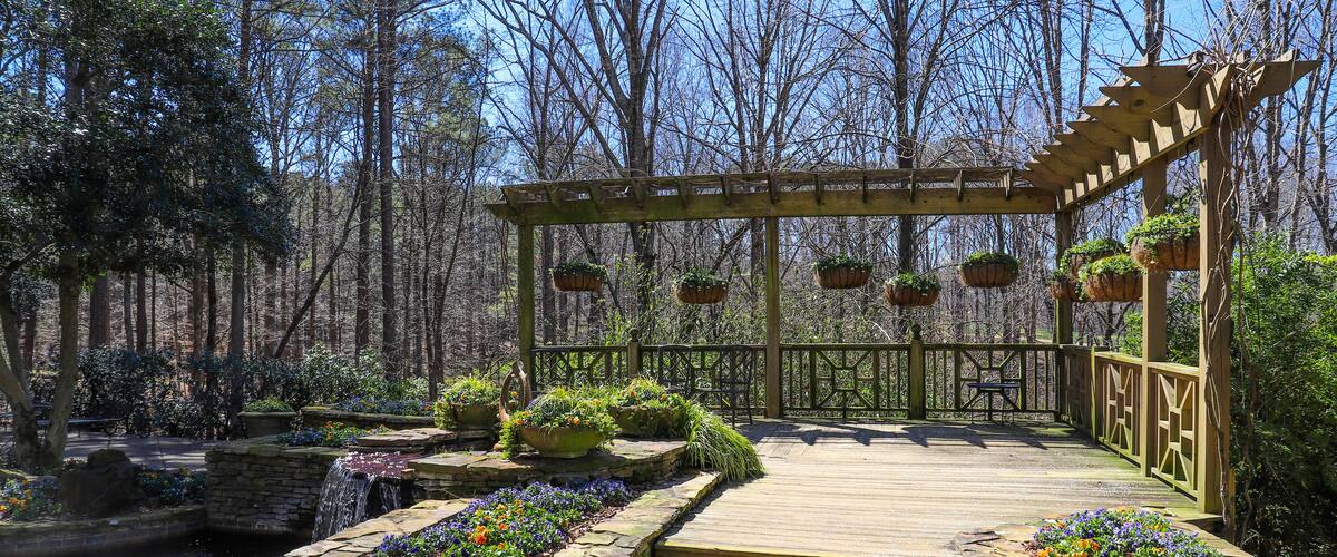 a gorgeous shot of a patio around a pool with a water fall surrounded by a brown wooden awning with plants hanging from it with colorful flowers and lush green trees and plants with blue sky
