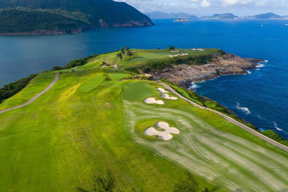 Un campo da golf costiero con un percorso sinuoso, bunker di sabbia e vista sull'oceano e sulle montagne lontane.