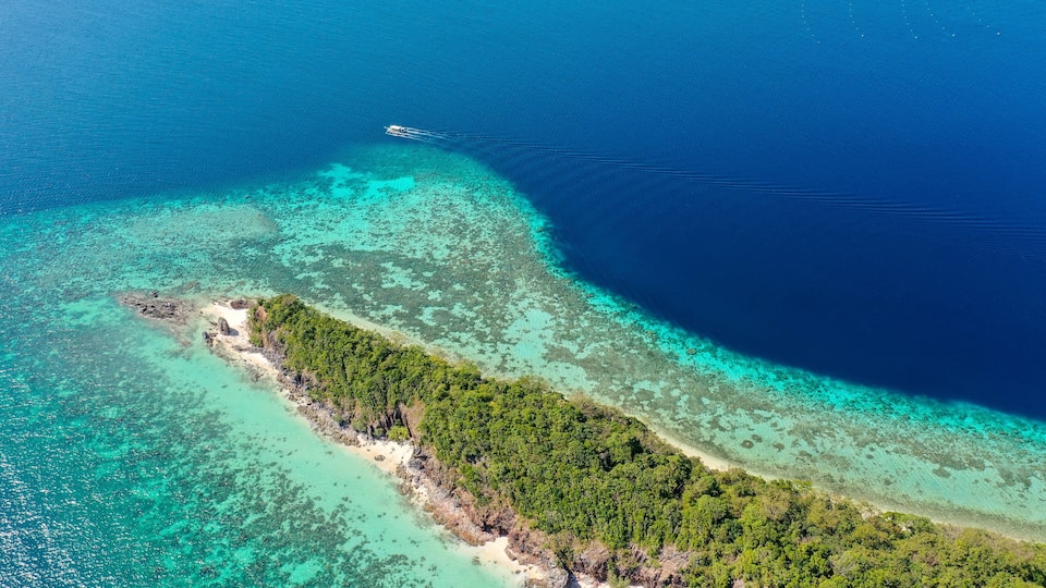 An aerial view of a tropical island with clear turquoise waters, a sandy beach, and dense green vegetation.