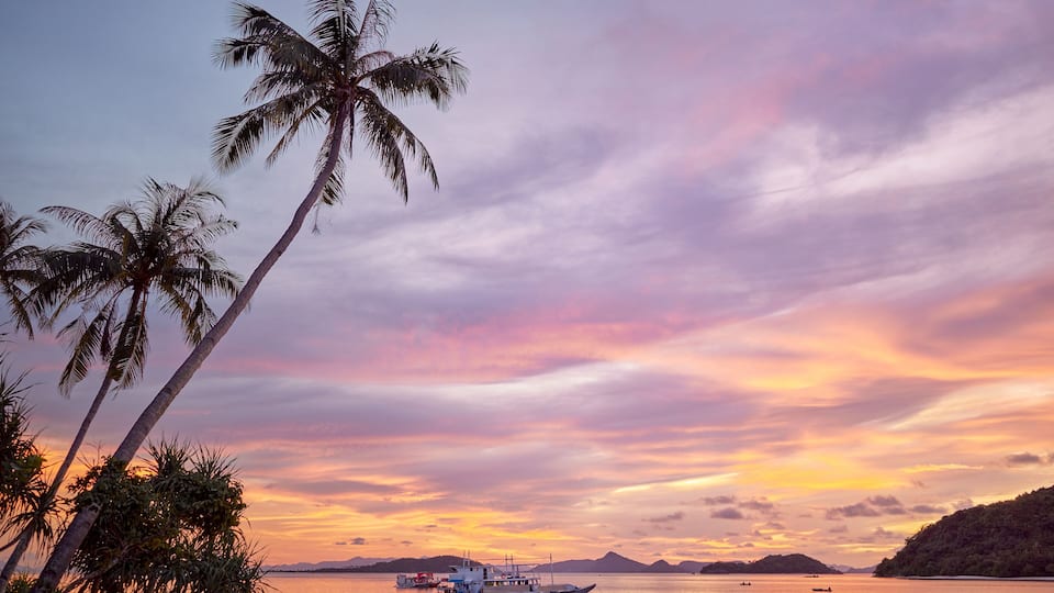 A sunset over a calm sea with palm trees and a boat.
