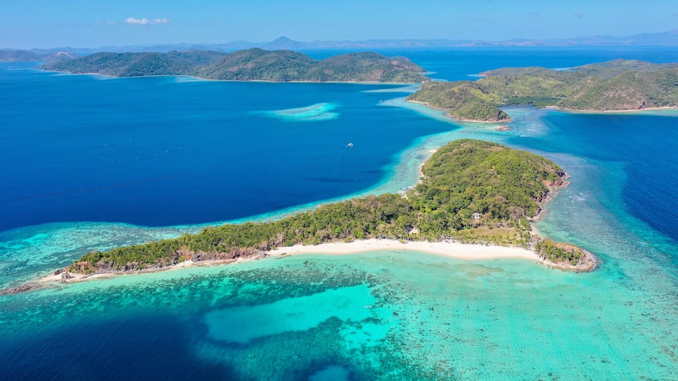 An aerial view of a tropical island with clear turquoise waters, surrounded by lush green hills and a small beach.