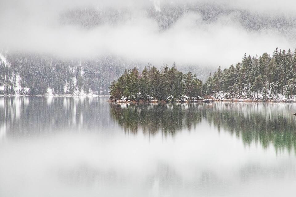 A calm lake with snow-covered trees and a misty forest in the background.