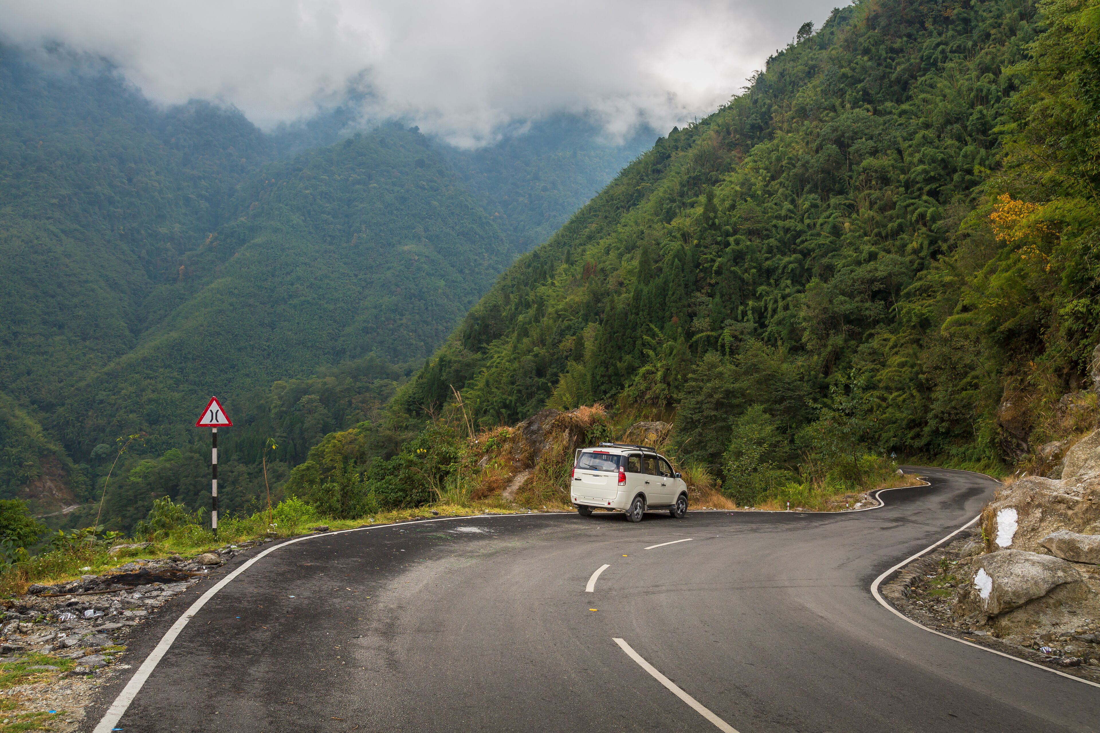 A winding mountain road with a van, surrounded by lush greenery and rocky terrain.