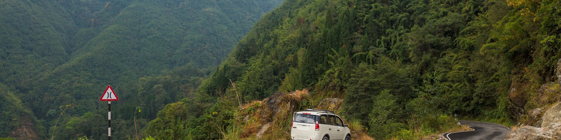 A winding mountain road with a van, surrounded by lush greenery and rocky terrain.