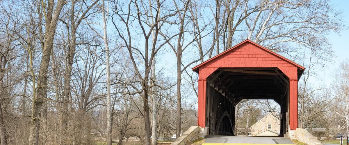A red covered bridge in a park with trees and a small building in the background.