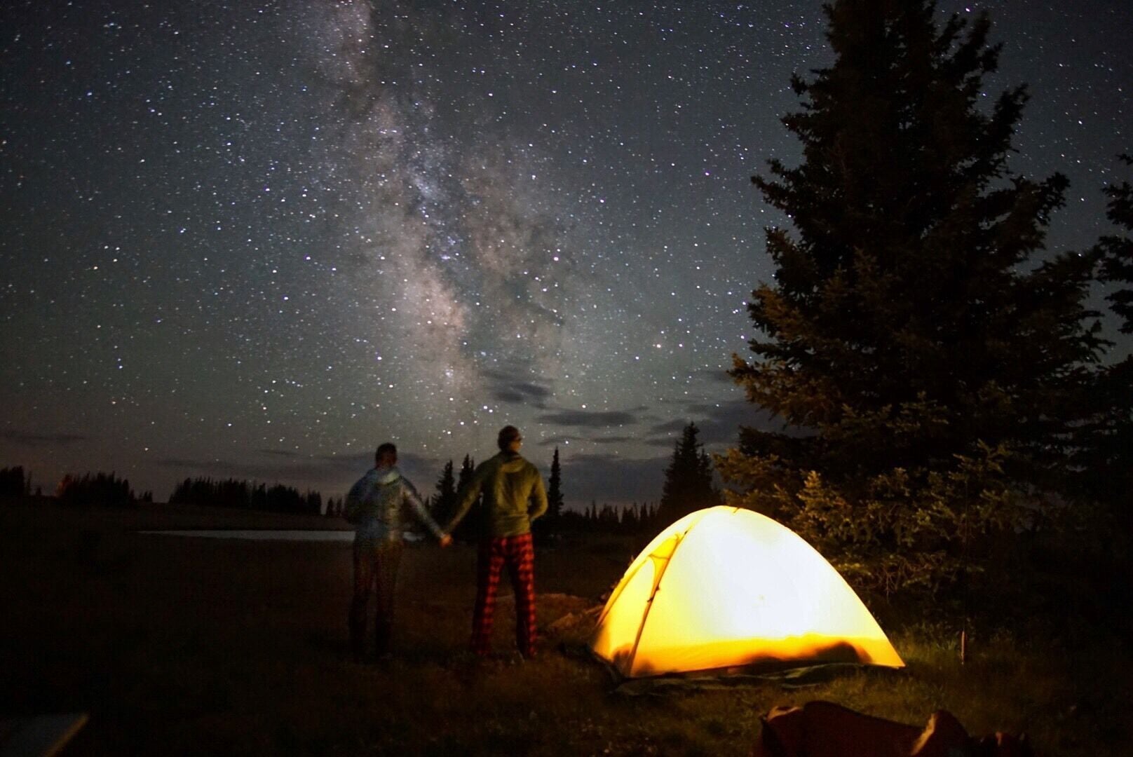 Two people holding hands under a starry sky with a tent in a forested area.