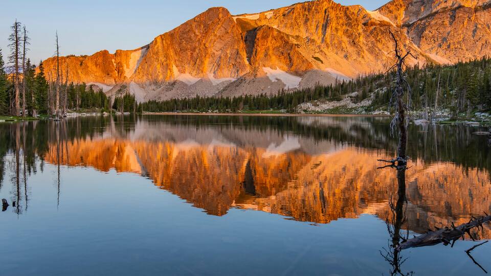A calm lake reflecting mountains, a clear sky, and a few trees.