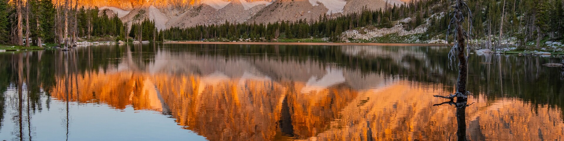Um lago tranquilo refletindo montanhas, céu claro e algumas árvores.