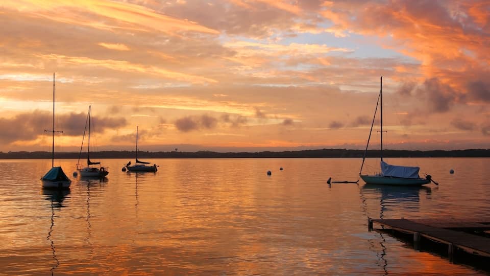 A calm lake at sunset with several sailboats anchored near the shore.