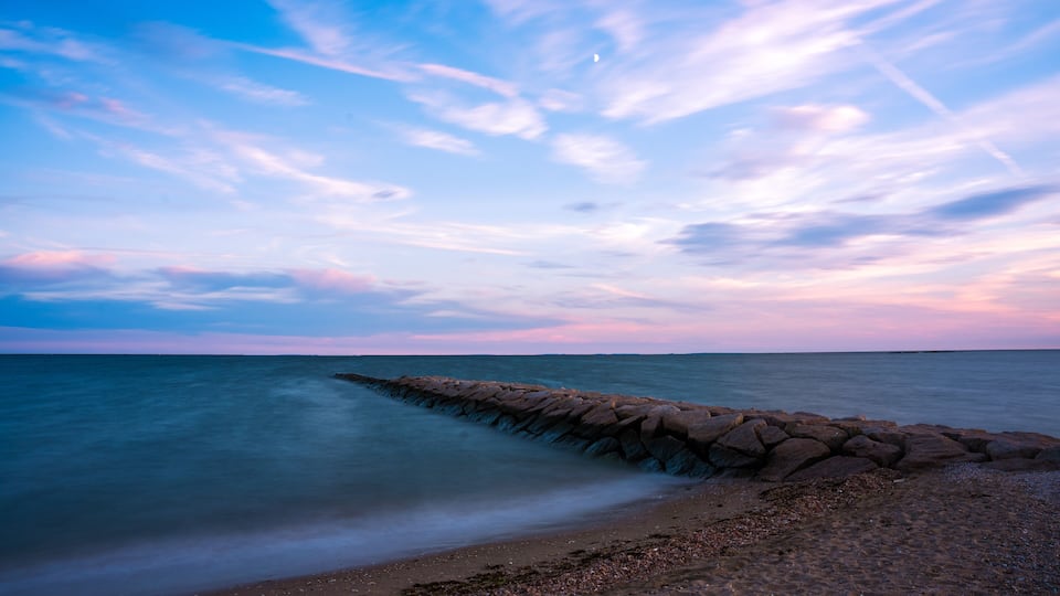 View of a stone jetty stretches into the tranquil ocean under a pastel sky brushed with pink and blue hues, captured from the sandy shore, Madison, Connecticut, United States.