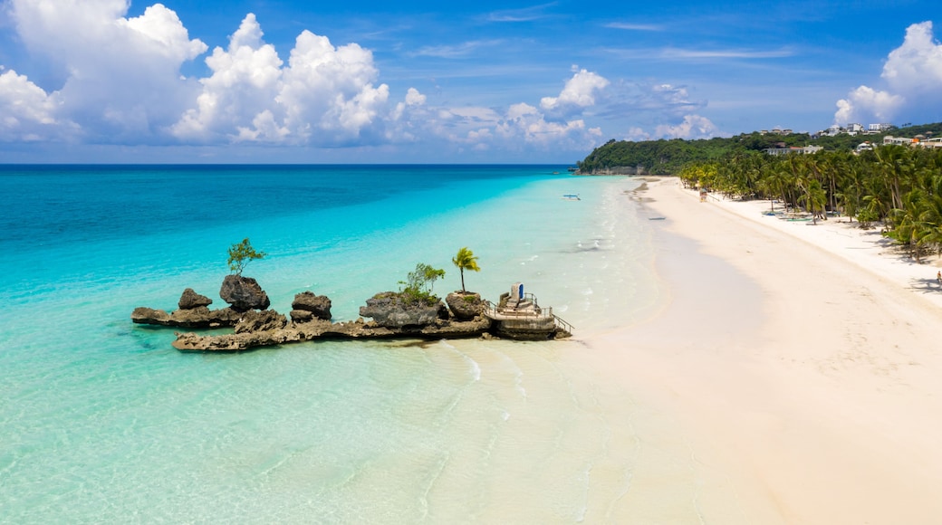 A tropical beach with clear turquoise waters, white sand, and a rocky outcrop with palm trees.