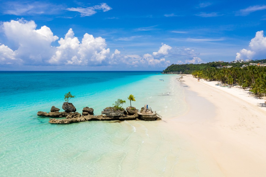 Una playa tropical con aguas turquesas cristalinas, arena blanca y un afloramiento rocoso con palmeras.
