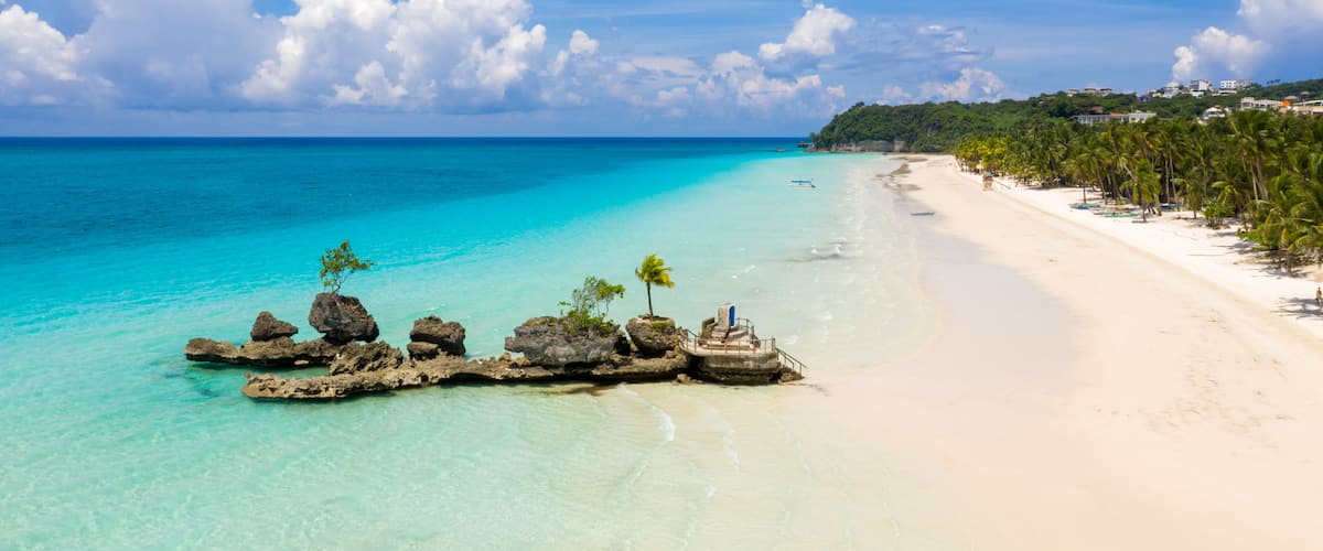 A tropical beach with clear turquoise waters, white sand, and a rocky outcrop with palm trees.