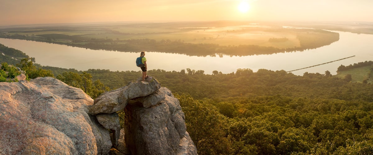Woman standing on sandstone overhang watching sunrise from summit of Petit Jean Mountain above Arkansas River ValleyÂ