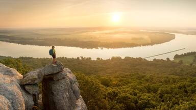Woman standing on sandstone overhang watching sunrise from summit of Petit Jean Mountain above Arkansas River ValleyÂ