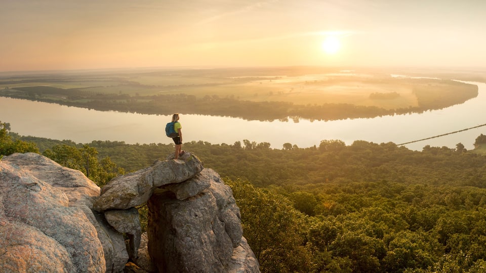 A person standing on a rock overlooking a river and forest during sunrise.