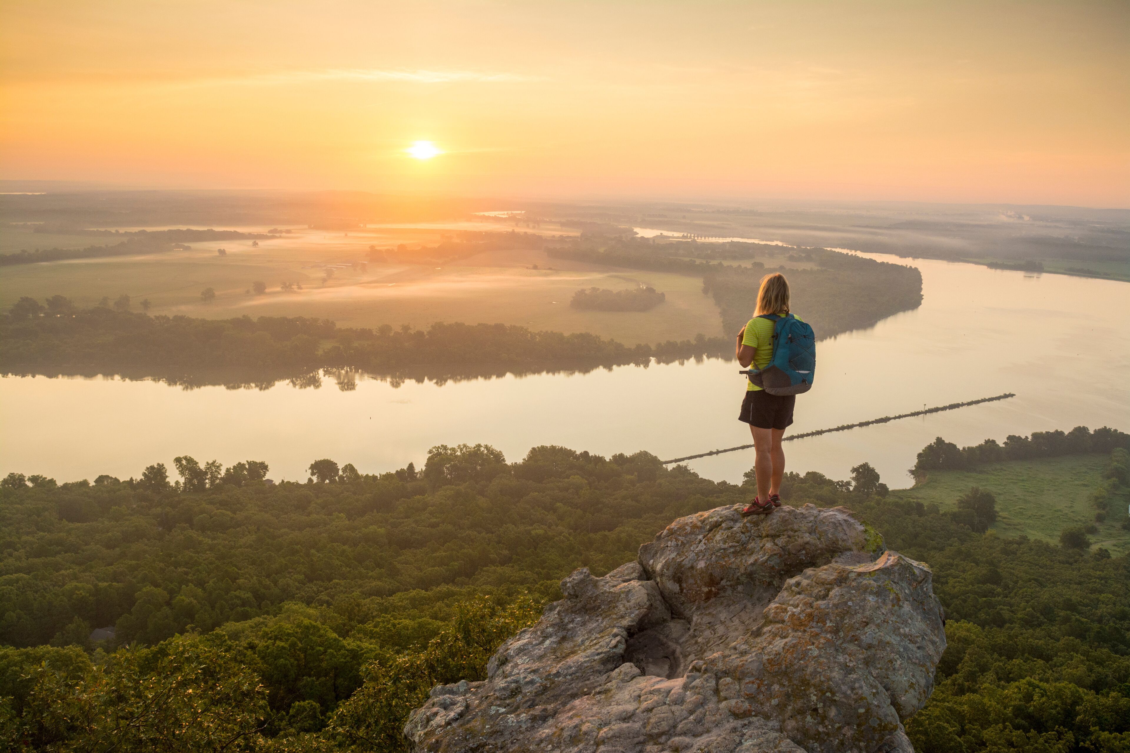 Woman standing on sandstone overhang watching sunrise from summitÂ ofÂ PetitÂ Jean MountainÂ above Arkansas River ValleyÂ 