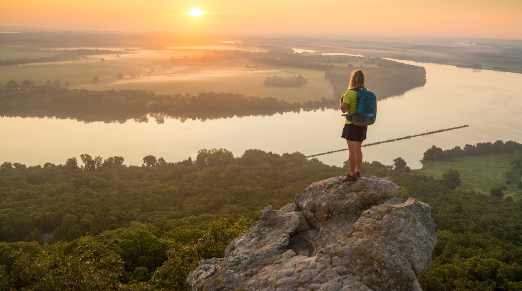 Woman standing on sandstone overhang watching sunrise from summit of Petit Jean Mountain above Arkansas River ValleyÂ