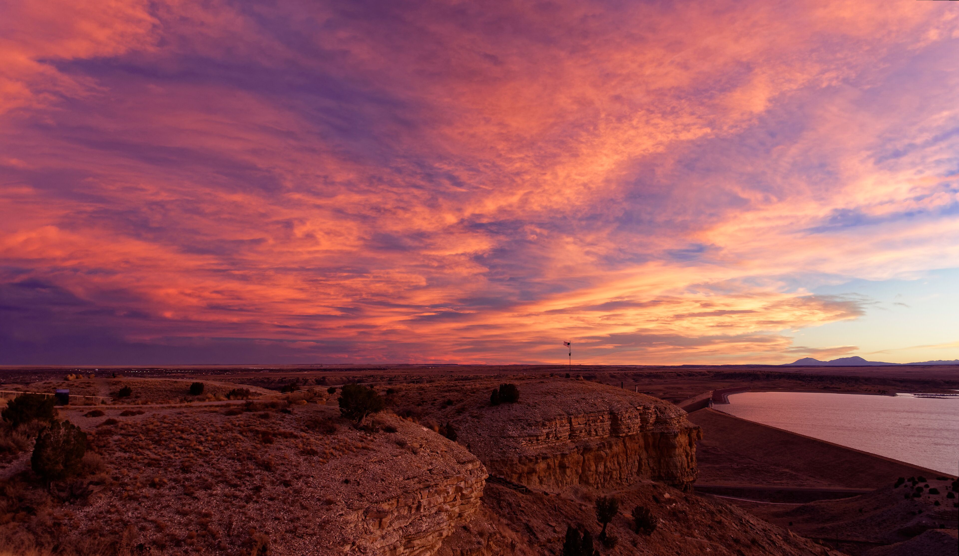 A desert landscape with a body of water, rocky formations, and a sunset sky.